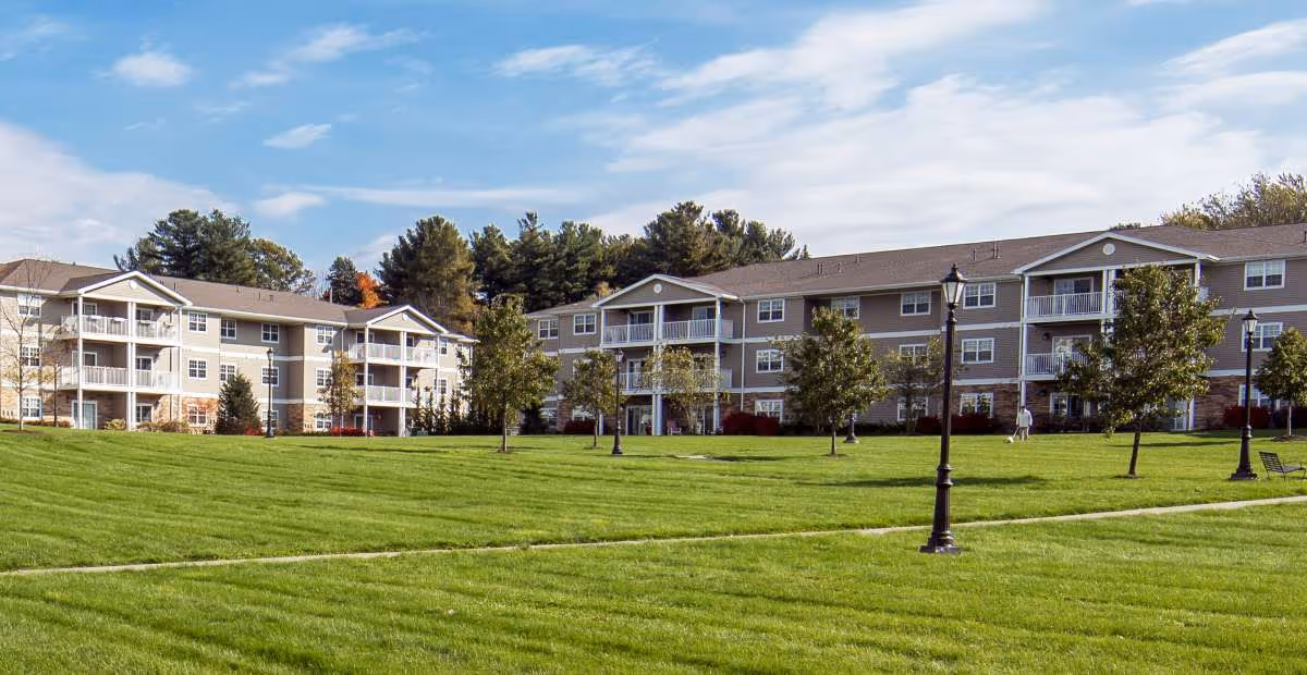 Exterior view of a senior living facility named Summit at Eastwyck, showing two three-story buildings with balconies, surrounded by a well-maintained green lawn, trees, and lampposts under a partly cloudy sky.