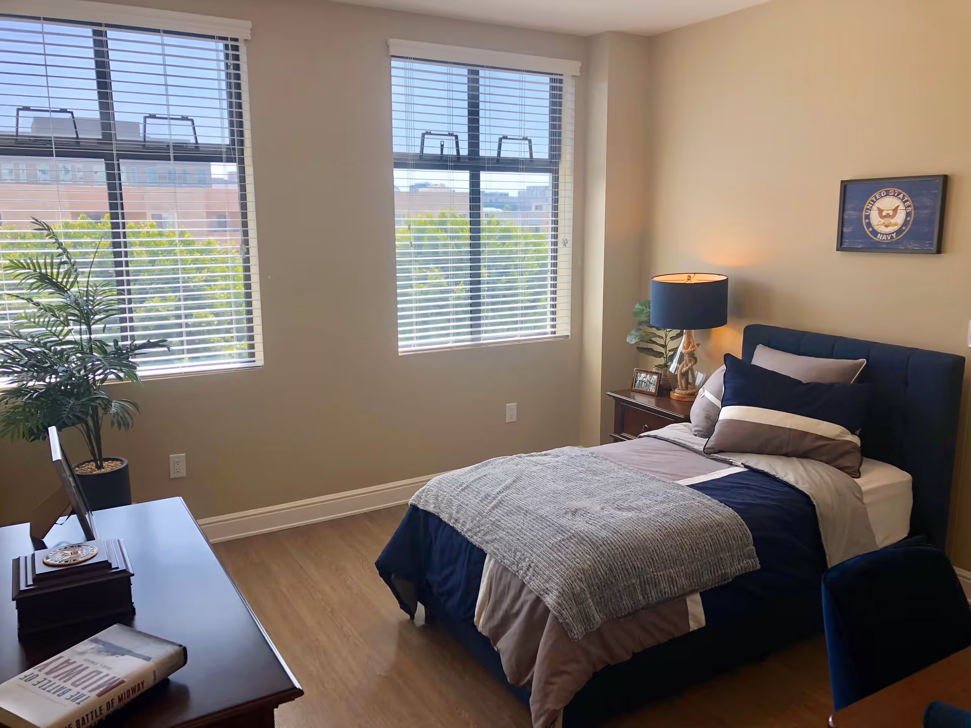A bright bedroom with two large windows with blinds, a single bed with navy blue and gray bedding, a nightstand with a lamp and a small plant, a framed United States Navy emblem on the wall, a wooden dresser with a book and decorative items, and a potted plant near the window.
