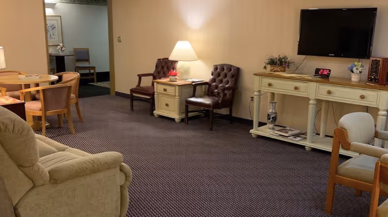 A cozy living room area with beige walls and carpeted floor. The room features a beige recliner chair, a round wooden dining table with four chairs, two dark brown leather chairs next to a small side table with a lamp, and a cream-colored console table holding a flat-screen TV, a clock, and decorative items.