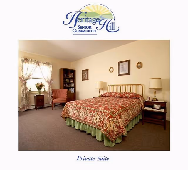 A cozy private suite bedroom in Heritage Hill Senior Community featuring a bed with a floral patterned bedspread and green bed skirt, two bedside tables with lamps, a wooden bookshelf, a pink upholstered chair, and a window with floral curtains letting in natural light.