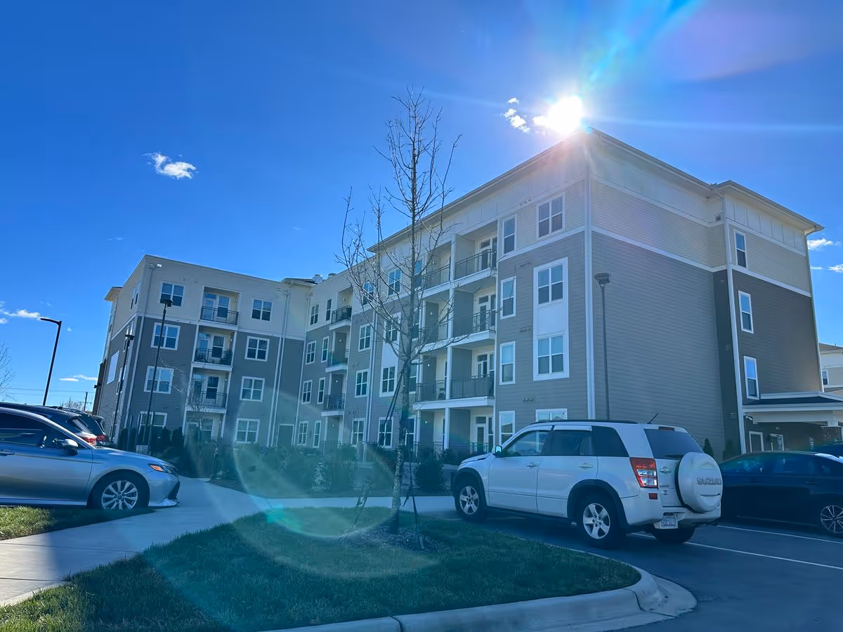 Four-story apartment building with balconies, parked cars in the lot, and the sun shining in a clear blue sky.