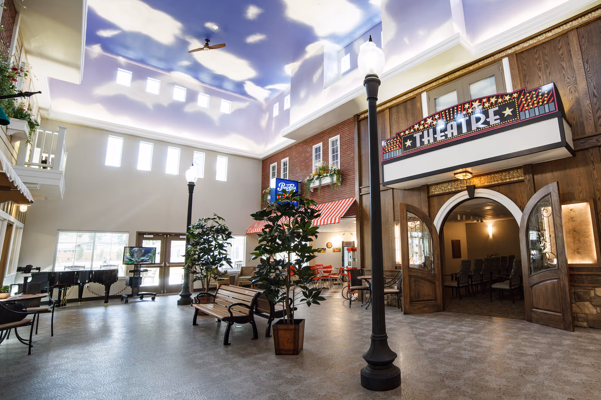 Interior of a senior living facility with a high ceiling painted to resemble a blue sky with clouds. The space includes a theatre entrance with double wooden doors and a marquee sign reading 'THEATRE'. There are benches, potted plants, a black grand piano, and streetlamp-style lights. The area also features a small cafe with red chairs and tables, and windows letting in natural light.