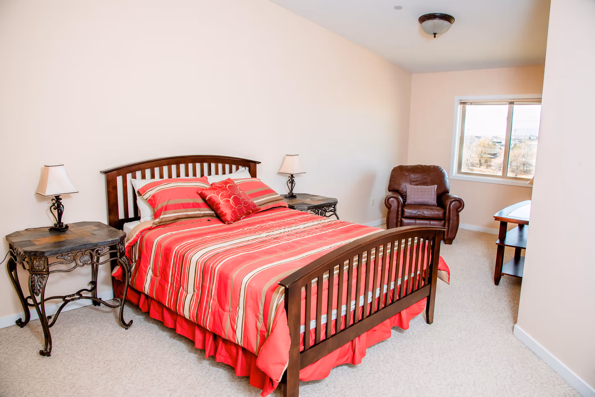 A bedroom with a wooden bed frame and a red striped bedspread. There are two matching bedside tables with lamps on each side of the bed. In the corner near a window, there is a brown leather armchair with a small pillow. A wooden table is also visible near the window. The walls are light-colored and the floor is carpeted.