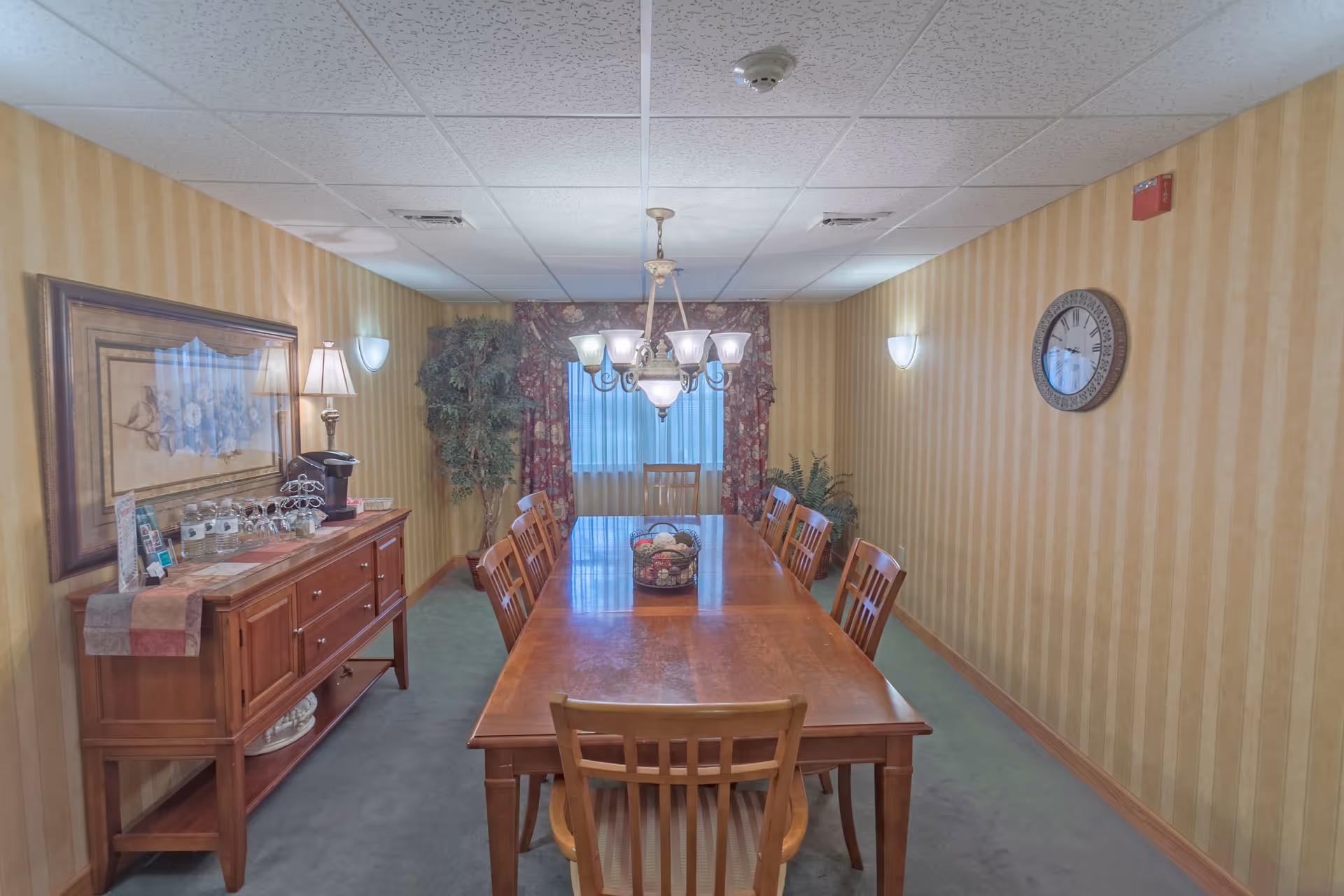 A dining room with a long wooden table surrounded by eight wooden chairs. A chandelier with multiple lights hangs above the table. On the left side, there is a wooden sideboard with a lamp, a coffee maker, and some glassware. The walls have yellow striped wallpaper, and there is a large framed picture on the left wall and a round clock on the right wall. Two potted plants are placed near the window at the far end of the room, which is covered with curtains.