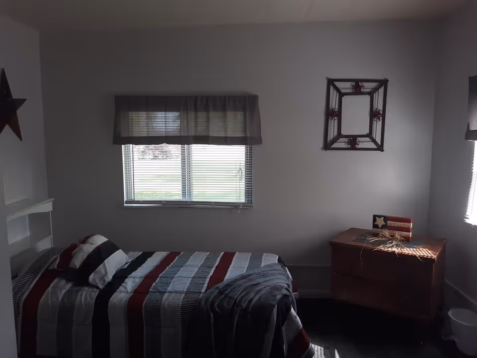 A small bedroom with a single bed covered in a striped quilt with red, white, gray, and black colors. There is a pillow and a folded blanket on the bed. A window with blinds and a valance is on the wall behind the bed. To the right, there is a wooden dresser with a decorative item and a small American flag-themed decoration on top. The walls are plain and light-colored, and there is a decorative star on the left wall and a framed wall decoration above the dresser.