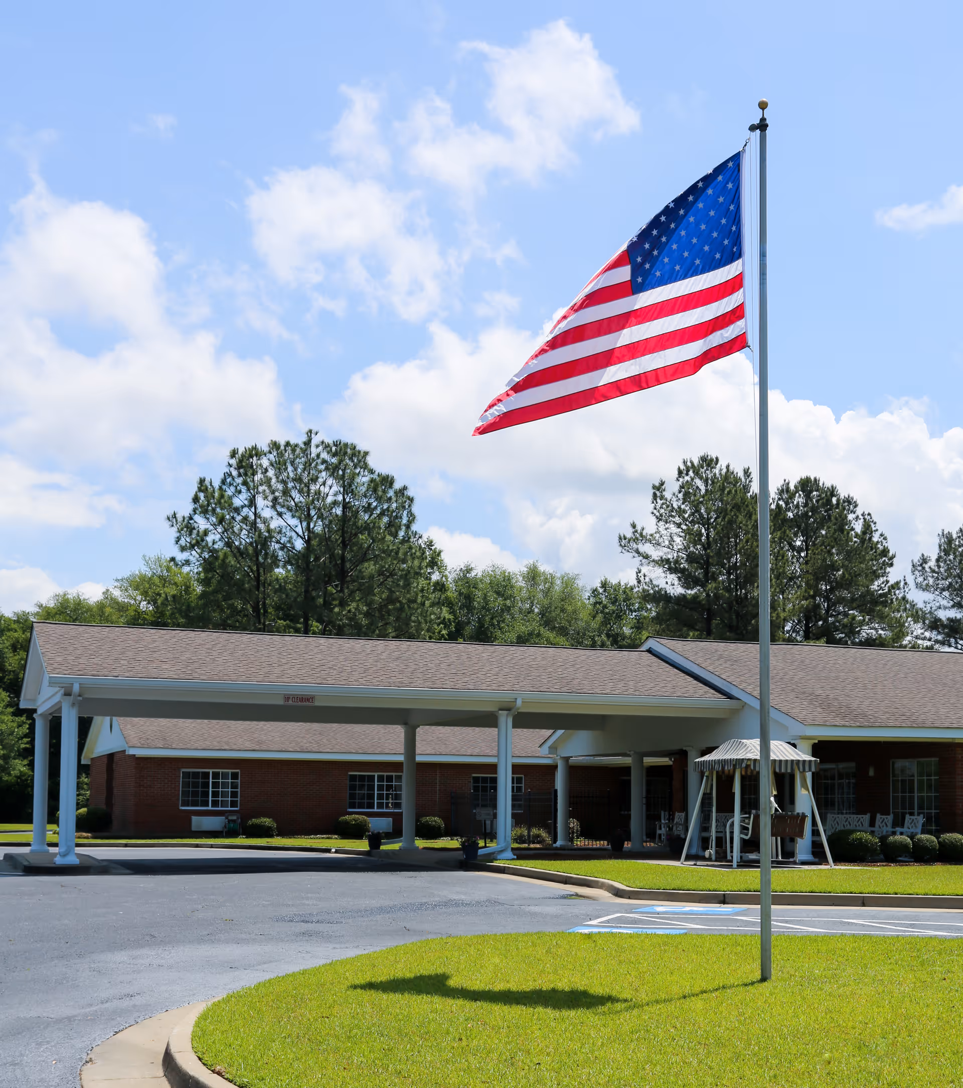 Exterior view of Lee County Health & Rehabilitation facility showing a single-story brick building with a covered entrance, an American flag on a flagpole in the foreground, green grass, and trees under a partly cloudy blue sky.