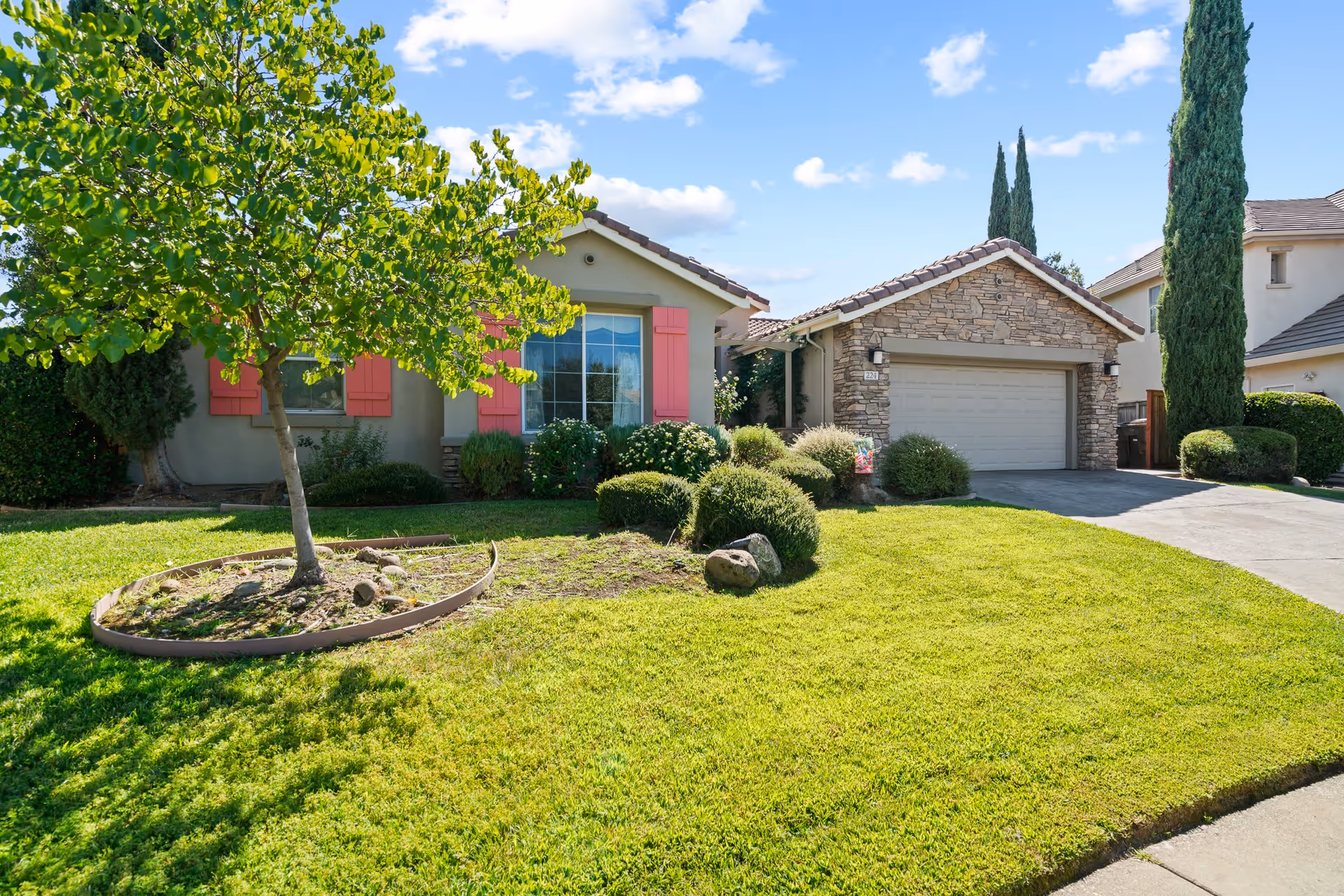 Front exterior view of a single-story house with a well-maintained lawn, a tree with green leaves, bushes, and a stone-faced garage. The house has beige walls with pink window shutters and a clear blue sky with some clouds in the background.