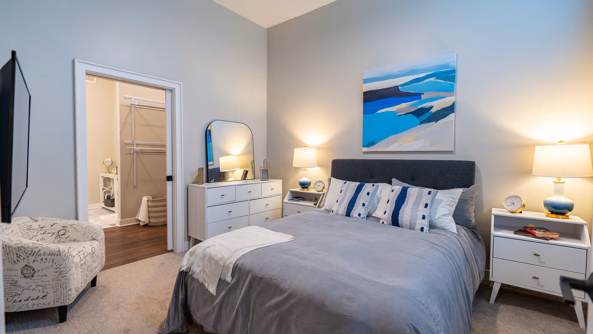 A modern bedroom with a gray upholstered bed featuring multiple decorative pillows and a gray bedspread. On either side of the bed are white nightstands with blue and white lamps and clocks. A white dresser with a large mirror stands against the wall near an open doorway leading to a walk-in closet and bathroom. A patterned armchair is positioned near the doorway. Above the bed hangs a large abstract painting in shades of blue and white.