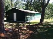 A dark shed sits beside a teal building in a grassy, tree shaded yard.
