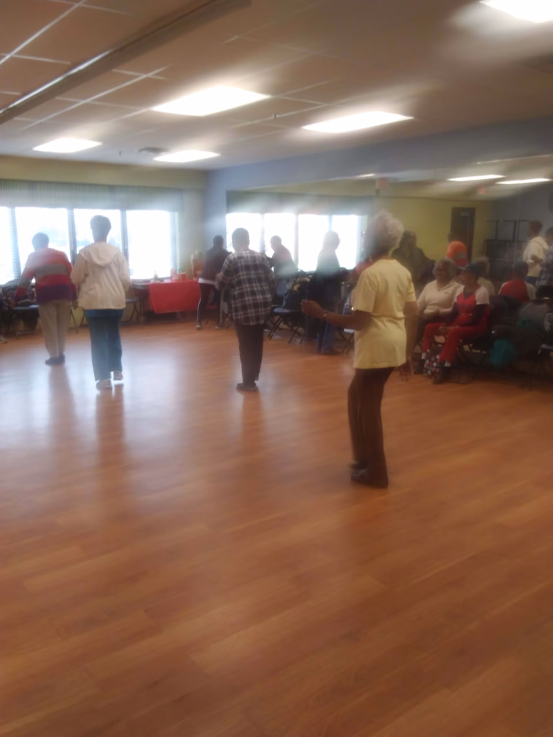 A group of elderly people standing and sitting in a spacious room with wooden flooring and large windows. Some individuals are standing and appear to be engaged in an activity, while others are seated along the wall. The room is well-lit with ceiling lights and has a mirror on one wall.