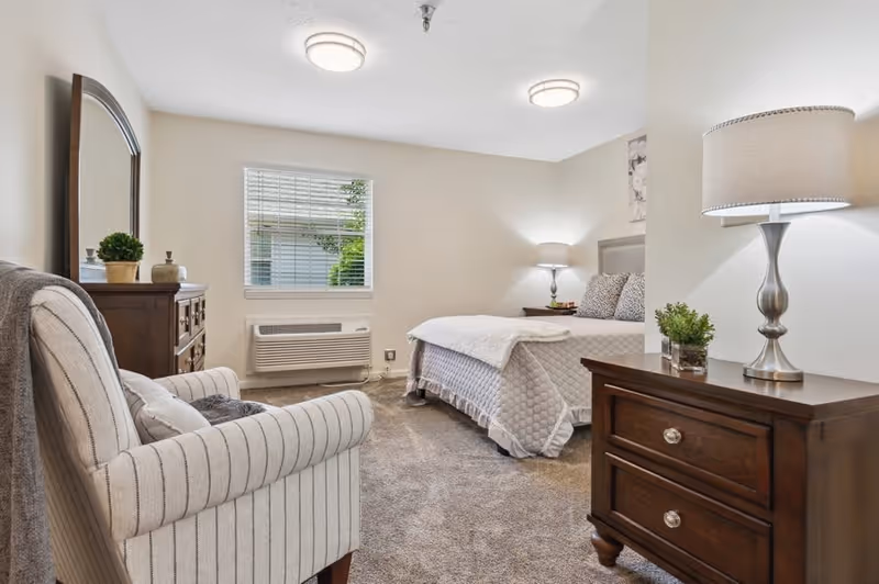 A cozy bedroom in a senior living facility featuring a bed with a quilted bedspread and pillows, a wooden nightstand with a lamp, a wooden dresser with a lamp and small plant, a striped upholstered armchair with a pillow and throw, a window with blinds, and beige carpeted floor.