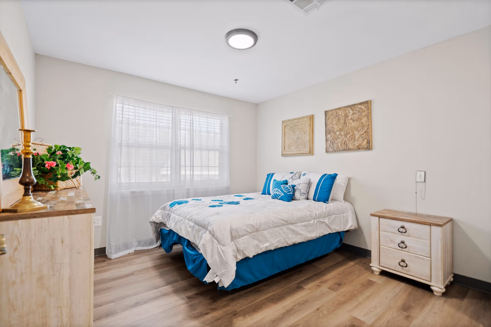 A bright and clean bedroom with a bed dressed in white and blue bedding, a wooden nightstand with three drawers, a dresser with a brass candle holder and a basket with flowers, two decorative wall hangings above the bed, and a large window with sheer white curtains allowing natural light to fill the room.