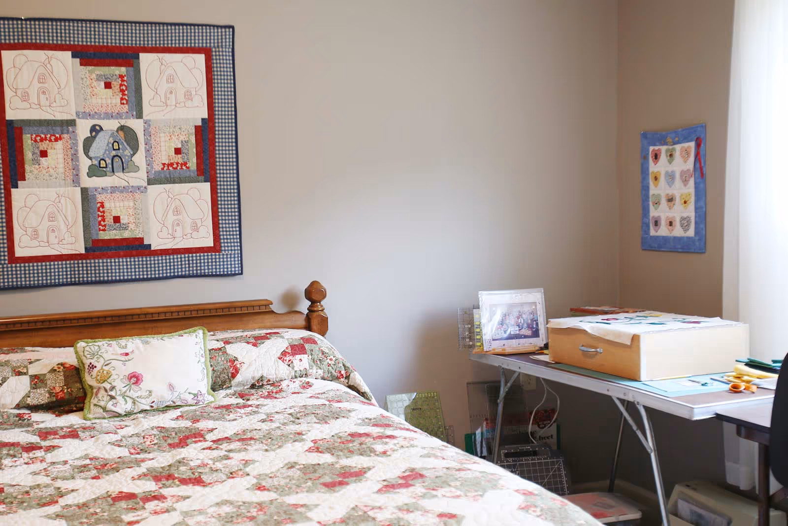 A cozy bedroom with a wooden bed dressed in a floral patchwork quilt, wall quilts, and a folding table holding crafting supplies.