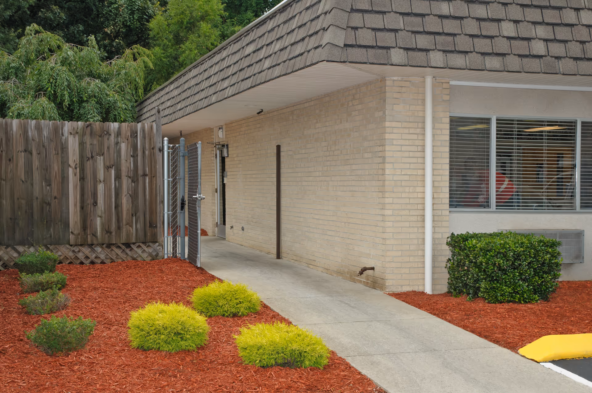 Outdoor walkway alongside a beige brick building with a wooden fence and metal gate on the left. The ground is covered with red mulch and small green bushes. A window with blinds is visible on the right side of the building.