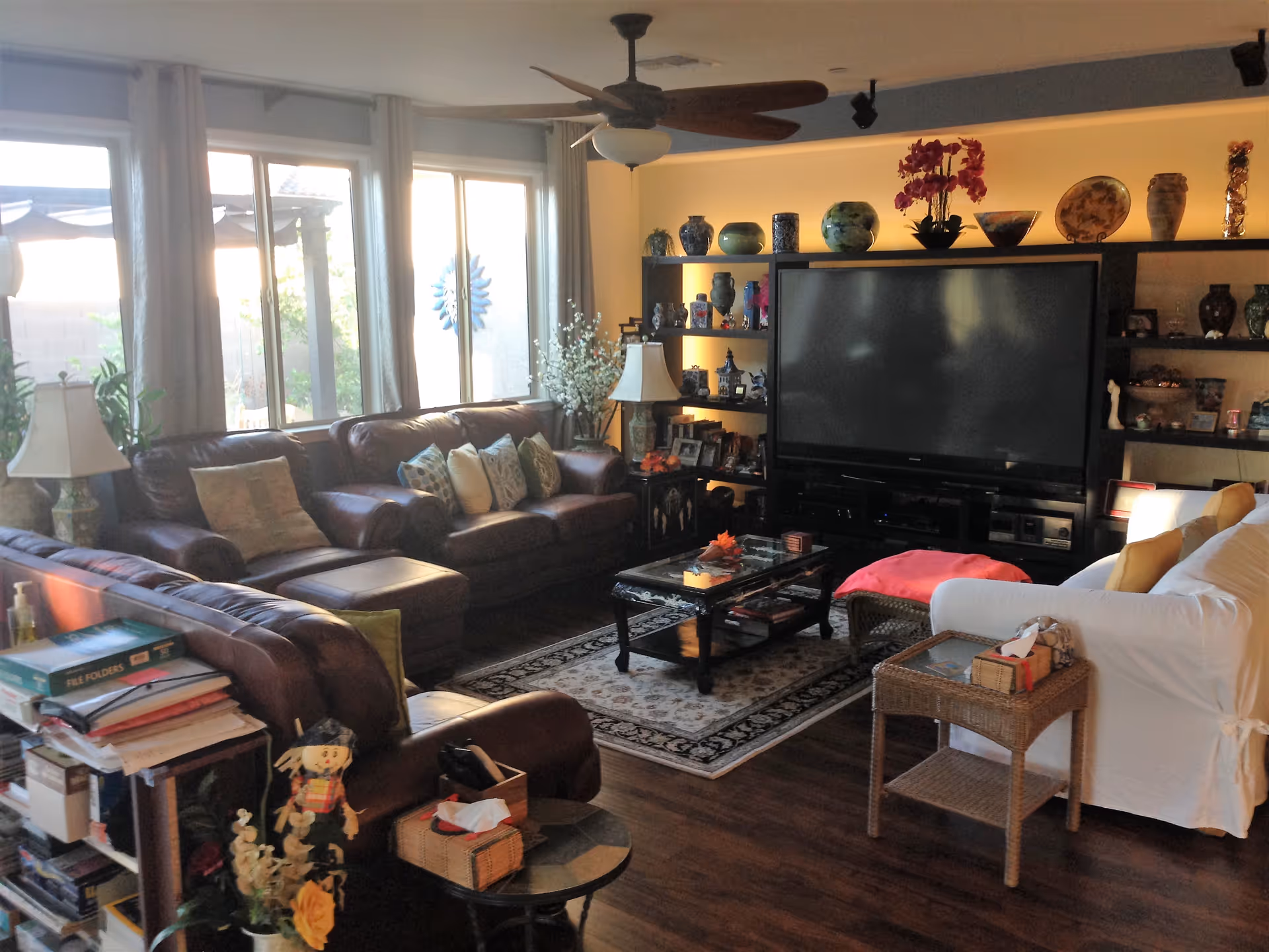 A cozy living room with large windows letting in natural light. The room features a dark brown leather sectional sofa with multiple cushions, a white armchair with yellow pillows, a glass coffee table on a patterned rug, and a large flat-screen TV on a black entertainment center filled with decorative vases and ornaments. There are two table lamps, a ceiling fan, and various decorative items including flowers and books.