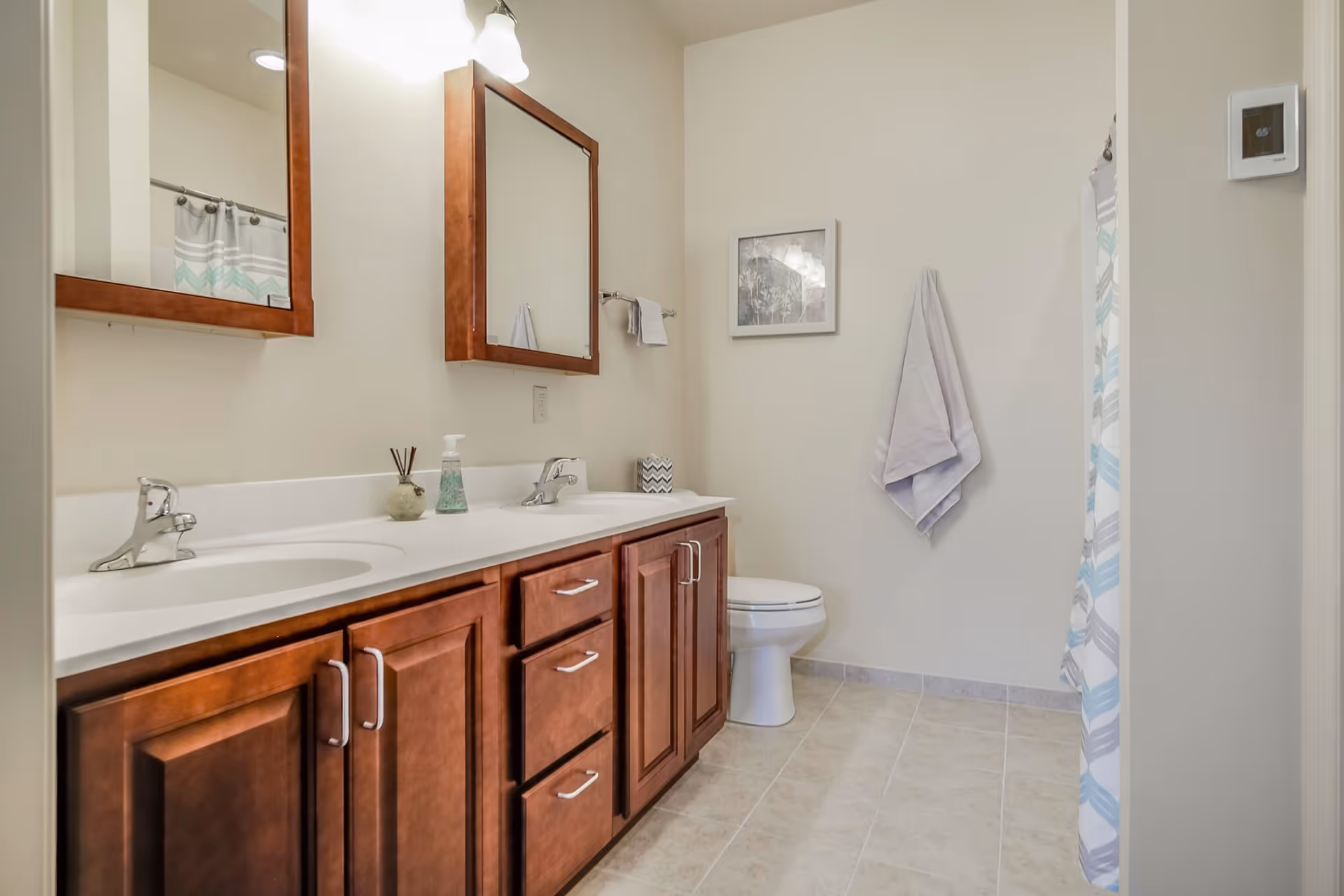 A clean bathroom featuring a double sink vanity with wooden cabinets and two mirrors above. There is a toilet next to the vanity, a towel hanging on the wall, a framed picture, and a shower with a patterned curtain. The floor is tiled and the walls are painted light beige.