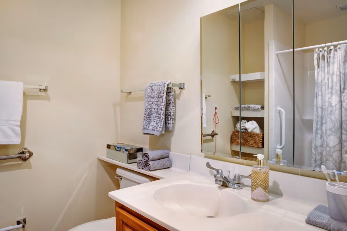Bright bathroom with a sink and mirror, folded towels, and a shower with a patterned curtain.