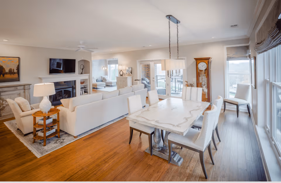 A bright and spacious living and dining area with a large white dining table surrounded by six upholstered chairs. The living room features a white sofa, armchairs, a fireplace with a mounted TV above it, and a side table with a lamp. Large windows on the right side let in natural light, and a grandfather clock stands near the glass doors leading to an outdoor patio.