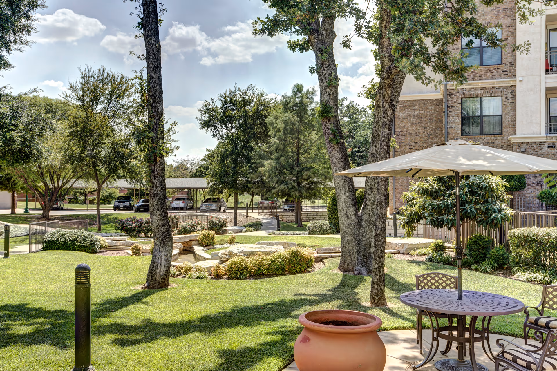 Outdoor garden area at Conservatory At Keller Town Center with green grass, trees, a round table with chairs and a large umbrella, a large terracotta pot, and a multi-story brick building in the background under a partly cloudy sky.