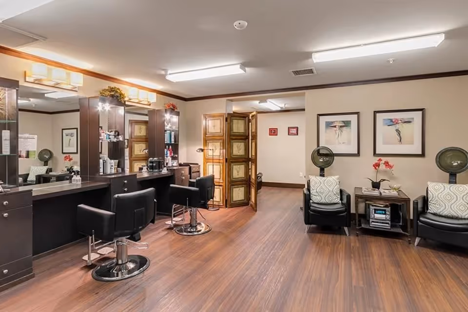 Well-lit salon with styling stations, black salon chairs, mirrors and hair dryers in a senior living facility.