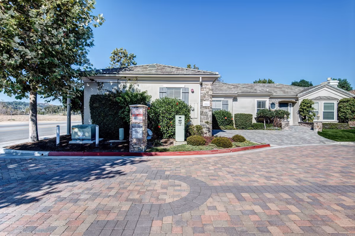 Exterior view of a single-story building with light-colored walls, stone accents, and a tiled roof. The building is surrounded by neatly trimmed bushes and trees. In front, there is a paved driveway and a red-painted curb with signs indicating no parking and fire lane. The sky is clear and blue.