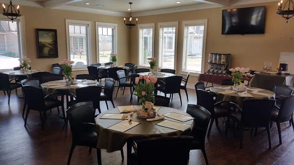 A dining room with multiple round tables covered with green tablecloths, each set with menus, napkins, and a vase of pink tulips. The room has large windows letting in natural light, a dark wood floor, and a wall-mounted TV above a beverage station with dispensers and cups.