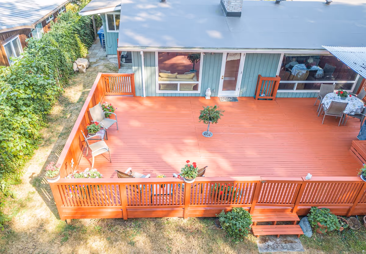 A spacious wooden deck attached to a light blue house with large windows and a door. The deck has several chairs, potted plants, a small table with a floral tablecloth, and a small tree in the center. The deck is surrounded by a wooden railing and steps leading down to a grassy yard with bushes along the fence.