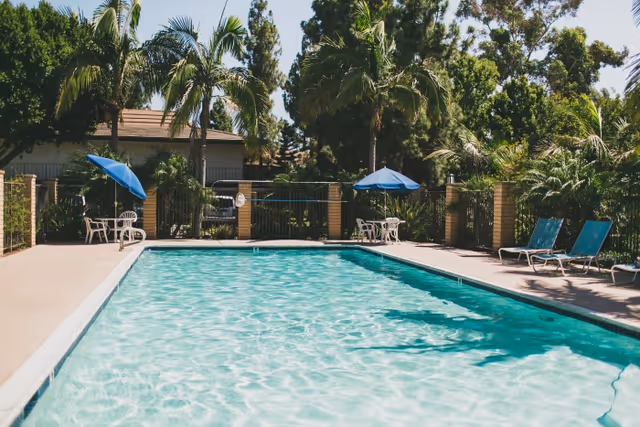 Outdoor swimming pool with lounge chairs and blue umbrellas surrounded by palm trees and a low building in the background.