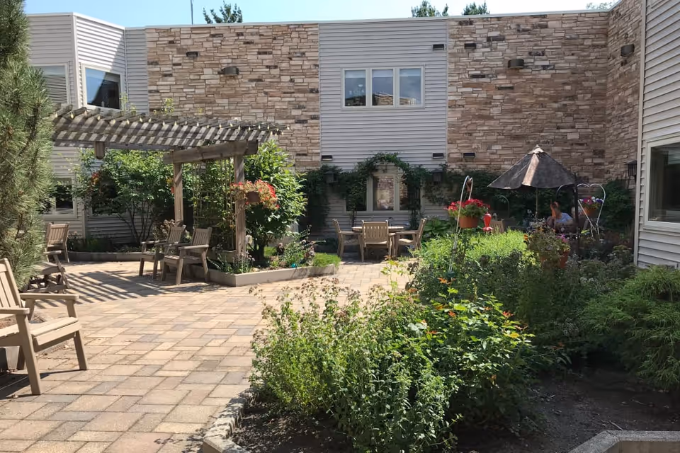 Outdoor courtyard area at Prairie Hills at Cedar Rapids featuring a paved patio with wooden chairs and tables, a wooden pergola, lush green plants, hanging flower baskets, and a person sitting under a large umbrella. The building exterior has stone and siding walls with windows overlooking the courtyard.