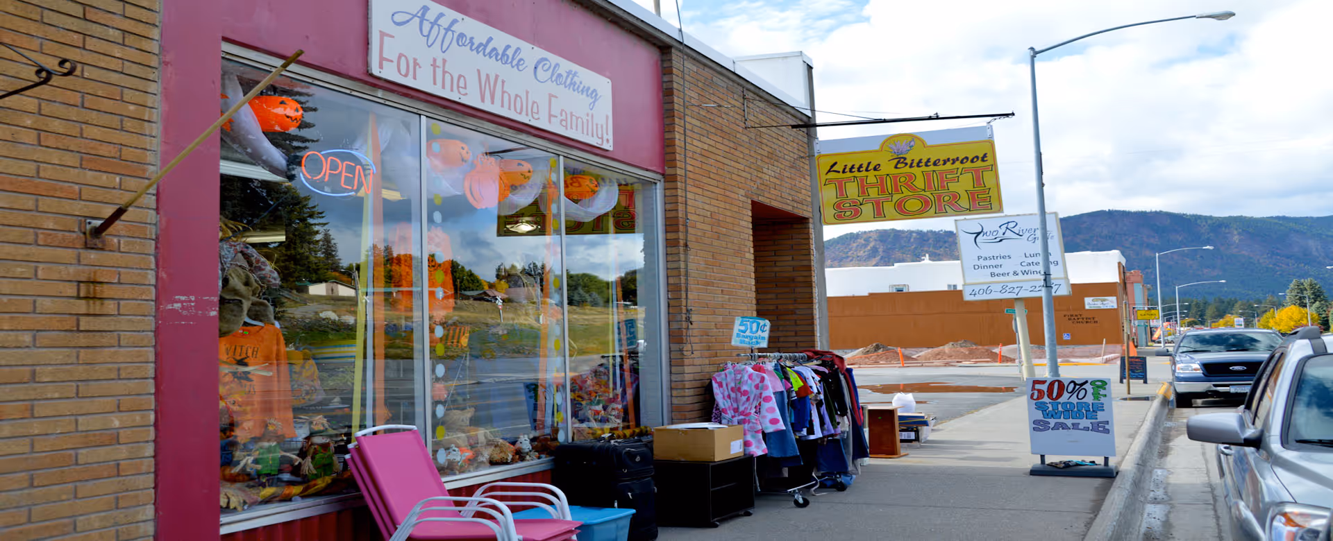Storefront thrift shop with clothing racks outside and a mountain backdrop.
