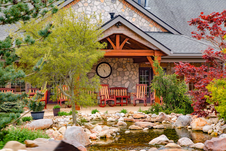 Outdoor patio area at The Inn at Walnut Trail featuring wooden rocking chairs and benches under a wooden pergola attached to a stone building. The foreground includes a landscaped garden with a small pond surrounded by rocks and various trees and shrubs with green and red foliage.