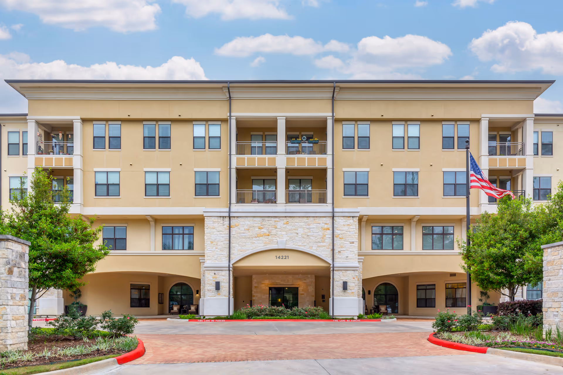 Front exterior of a multi-story senior living building with balconies, main entrance driveway, landscaping and an American flag.