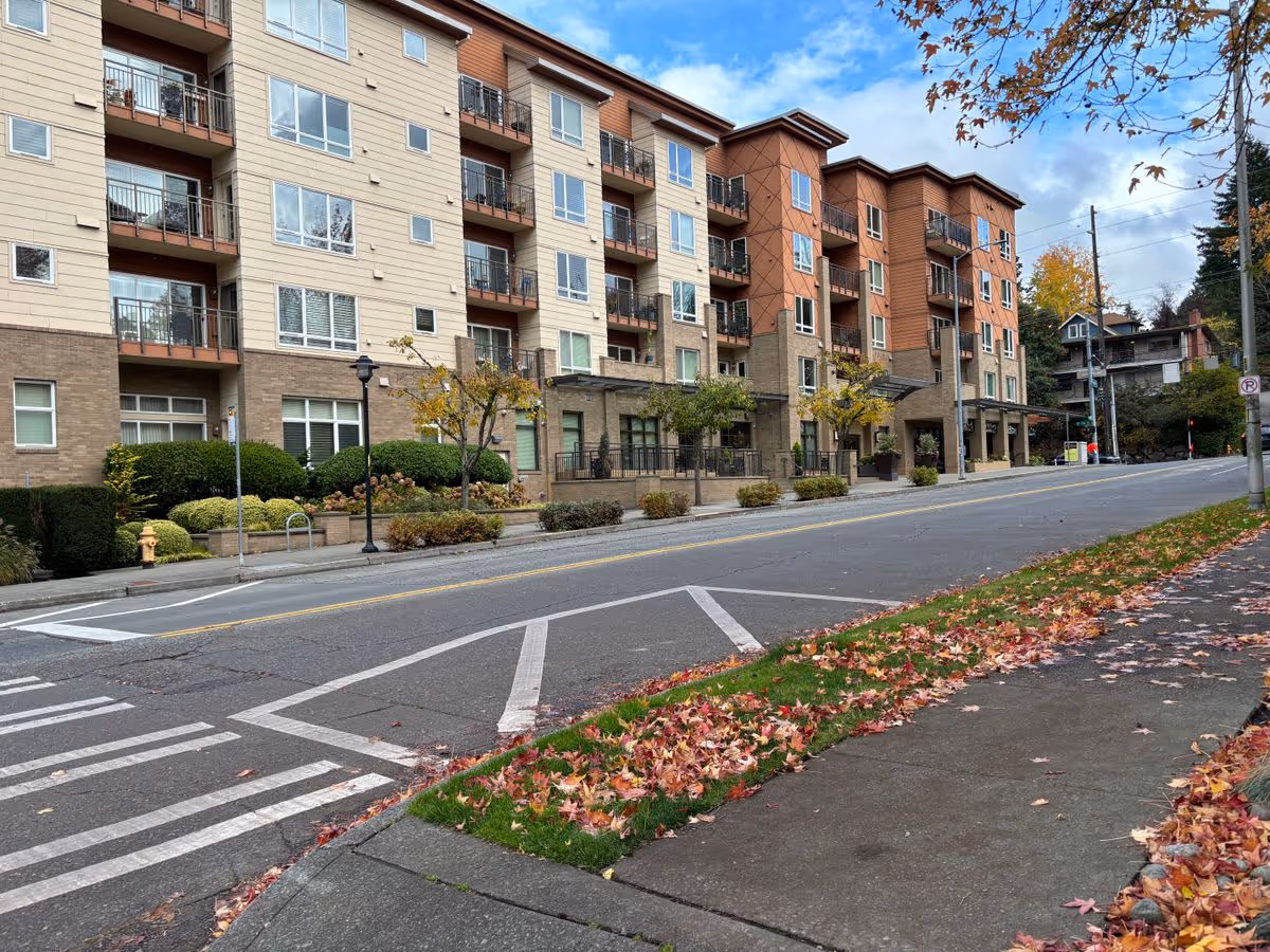 Street view of a multi-story senior living building with balconies, landscaped frontage, and autumn leaves along the sidewalk.