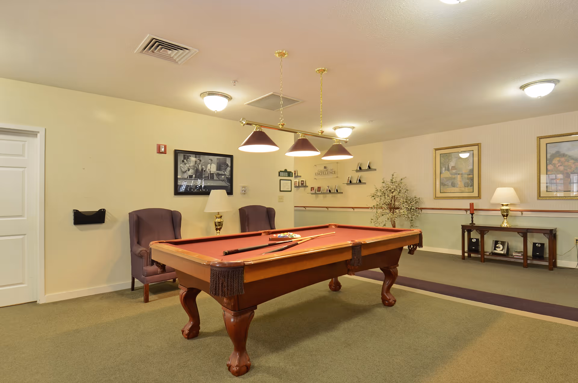 A senior living facility game room with a pool table in the center, two purple armchairs, a table lamp, framed pictures on the walls, and a small console table with decorative items and a lamp.