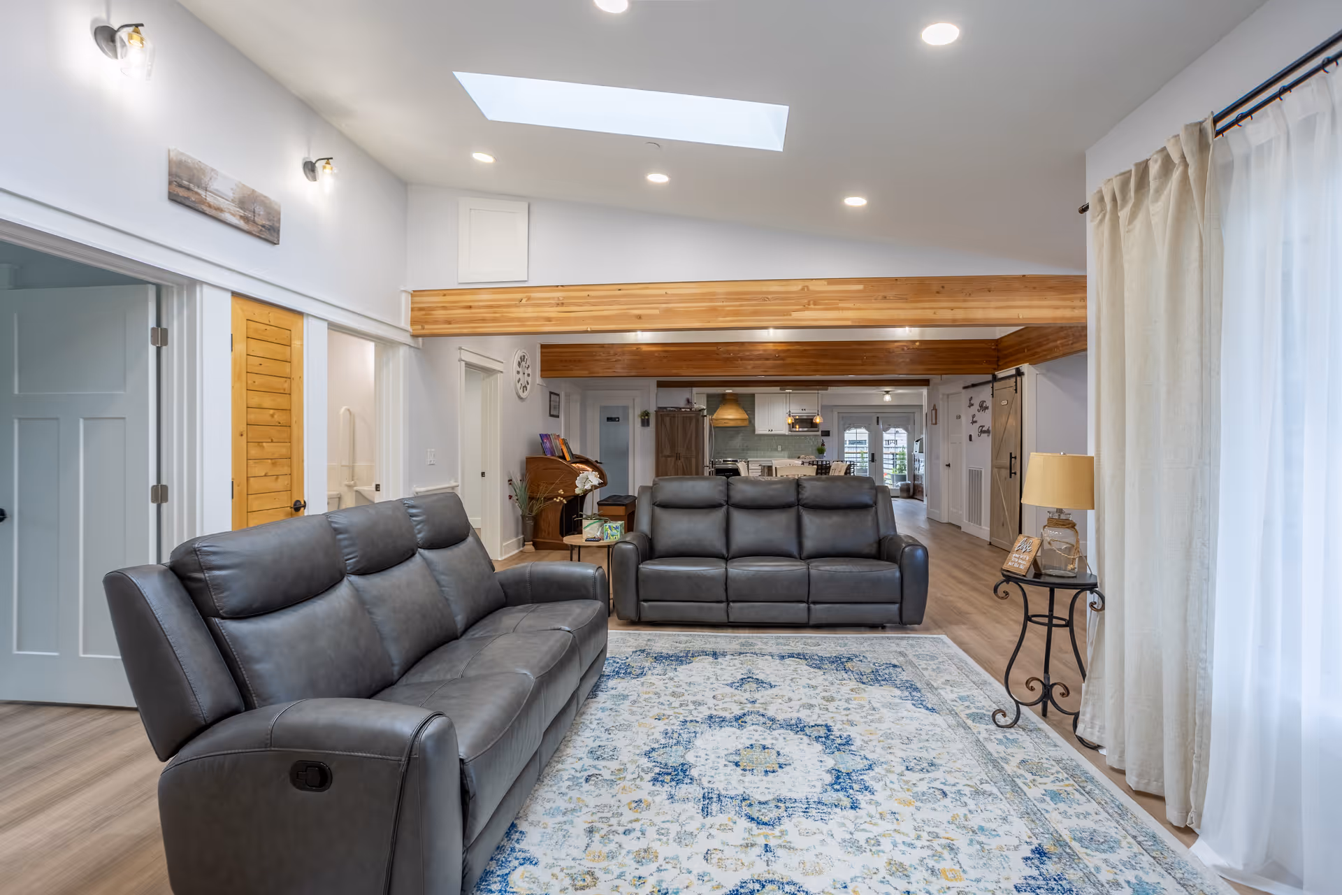 A spacious living room with two gray leather sofas facing each other on a patterned area rug. The room features wooden ceiling beams, a skylight, recessed lighting, and light-colored walls. To the right, there is a window with beige curtains and a small side table with a lamp. The background shows an open kitchen and dining area with wooden flooring.