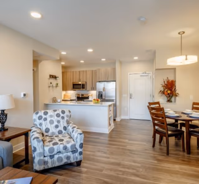 Open-plan living area with a patterned armchair, kitchen island with stainless appliances, and a dining table.