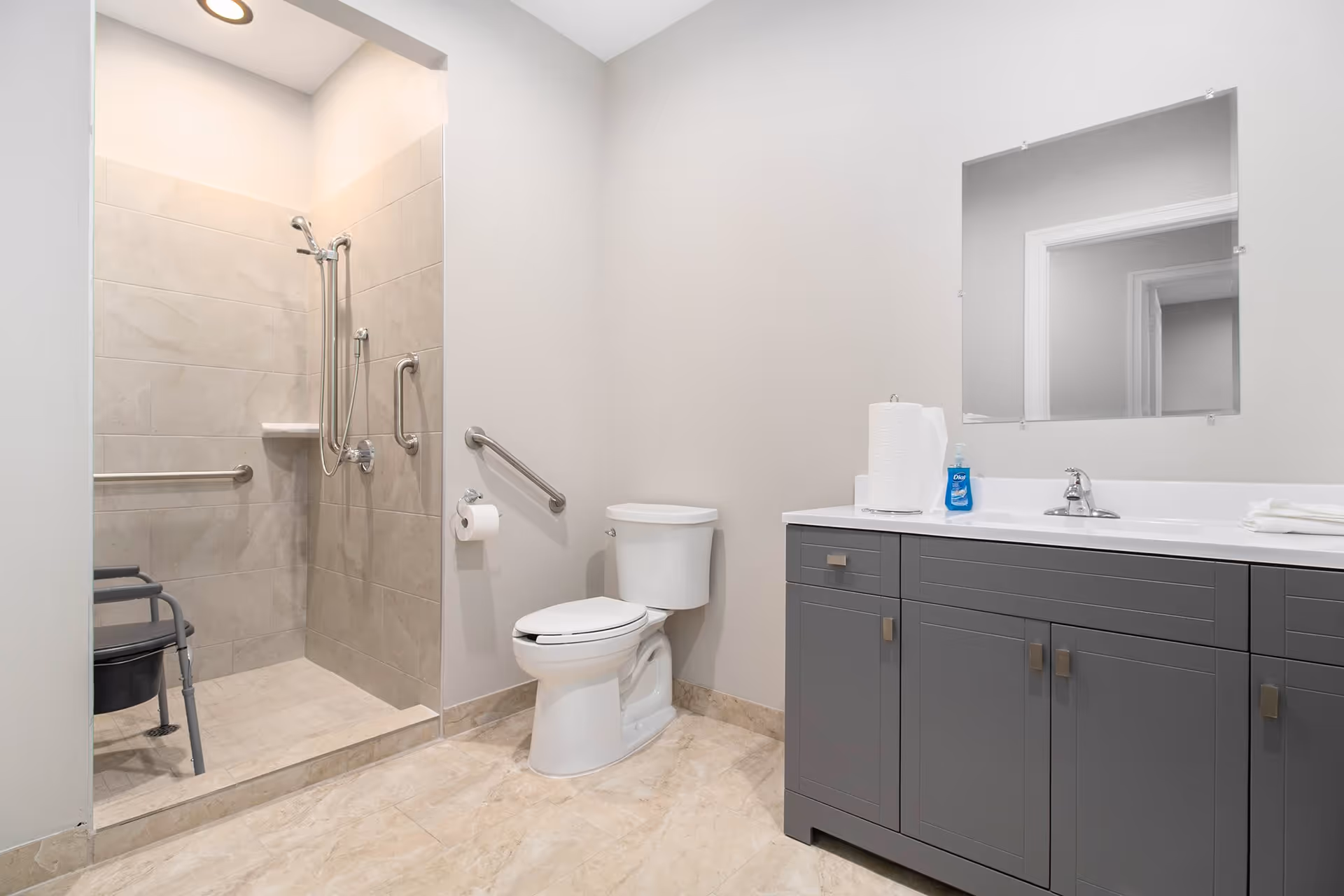 A clean and modern bathroom featuring a walk-in tiled shower with a shower chair and grab bars, a white toilet with a grab bar beside it, and a gray vanity with a white countertop, paper towels, hand soap, and a large mirror above.