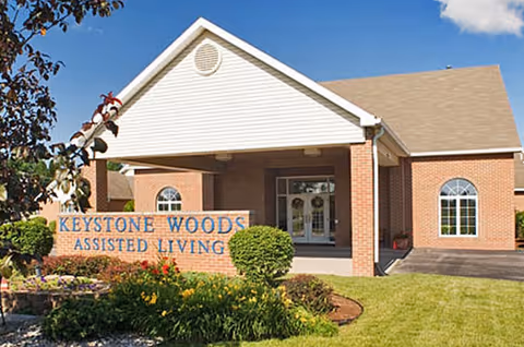 Front entrance of Keystone Woods Assisted Living building with a covered portico, sign, and landscaped lawn.