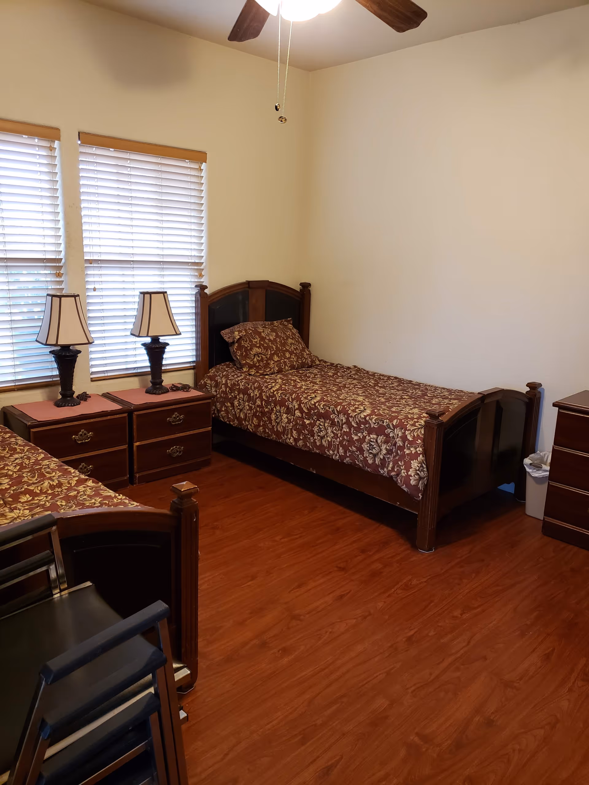 A bedroom with two single beds featuring dark wooden frames and floral patterned bedspreads. Between the beds are two wooden nightstands with matching table lamps. The room has wooden flooring, two windows with closed blinds, and a ceiling fan with a light fixture. There is also a small trash bin and a piece of furniture with drawers on the right side.