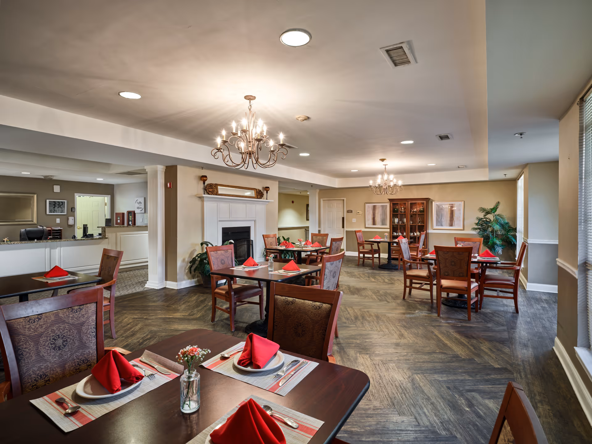 A spacious dining room in an assisted living facility with several wooden tables and chairs. Each table is set with white plates, silverware, and red folded napkins. The room features two ornate chandeliers, a fireplace with a decorative mirror above it, a wooden cabinet with glass doors, and large windows with blinds allowing natural light to enter. The floor has a wood-patterned design, and there are plants placed around the room for decoration.
