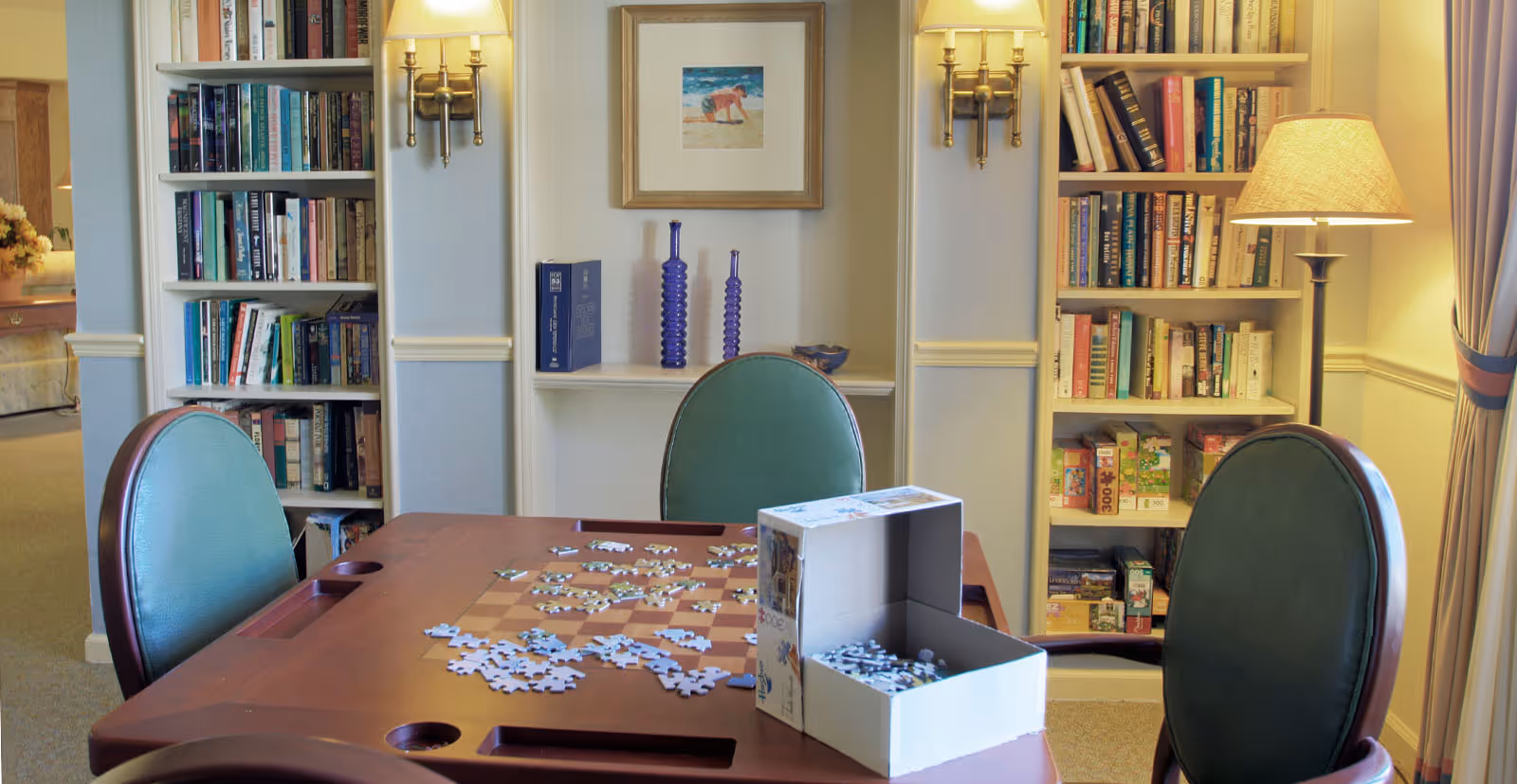 A cozy activity room with a table covered in jigsaw puzzle pieces, surrounded by green-upholstered chairs and bookshelves with lamps.