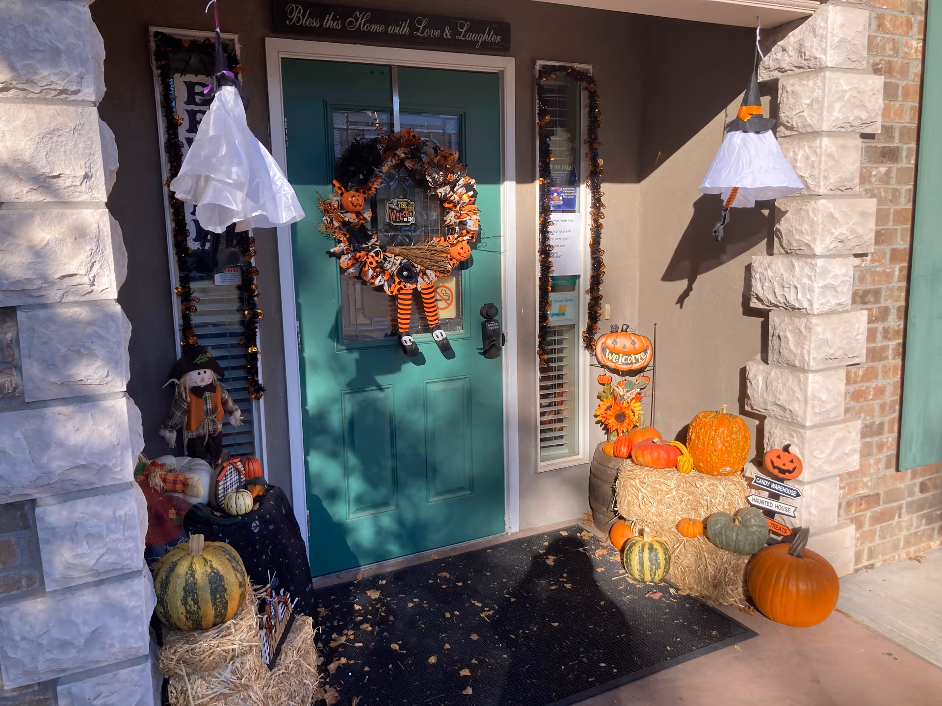 Entrance door decorated for Halloween with a wreath featuring orange and black elements and dangling striped legs. The doorway is flanked by two windows adorned with garlands. There are various pumpkins, hay bales, and Halloween-themed decorations including a small scarecrow and hanging ghost figures on either side of the door. A sign above the door reads 'Bless this Home with Love & Laughter.'
