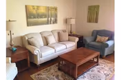 A cozy living room with a beige sofa, a blue armchair, a wooden coffee table, two side tables with lamps, and two framed pictures of trees on the walls.