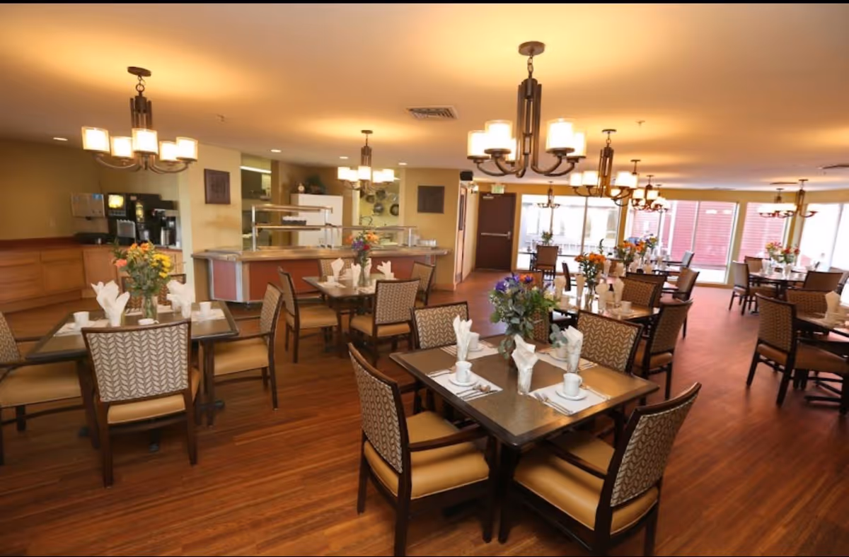 A spacious dining room in an assisted living facility with multiple tables set for meals. Each table has chairs with patterned backs and cushioned seats, white napkins folded elegantly, cups, and silverware. There are flower arrangements on each table. The room has warm lighting from multiple chandeliers and large windows letting in natural light. A serving counter and beverage station are visible in the background.
