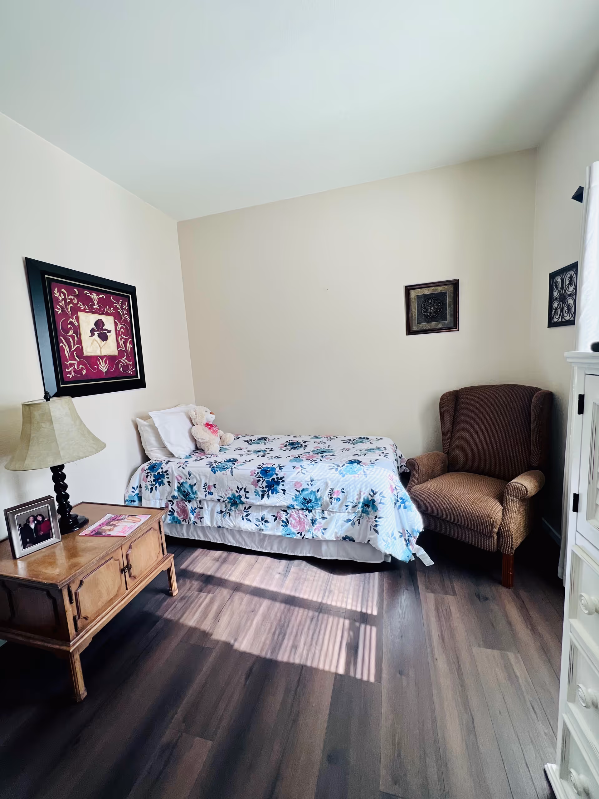 A small bedroom with a single bed covered in a floral bedspread, a teddy bear on the bed, a wooden nightstand with a lamp and framed photo, a brown upholstered armchair, and framed artwork on the walls. Sunlight streams in through a window, casting shadows on the wooden floor.