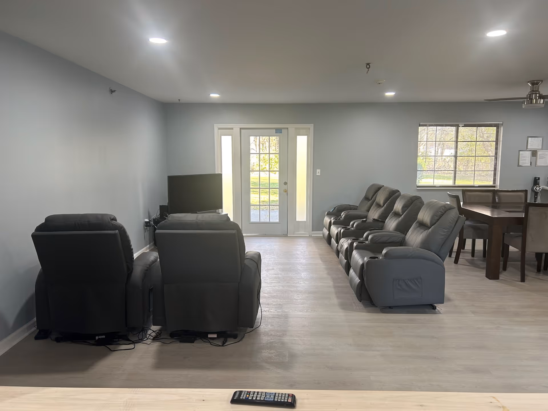 A spacious communal lounge with rows of gray recliners facing a TV, a dining table by a window, and a glass door at the far end.