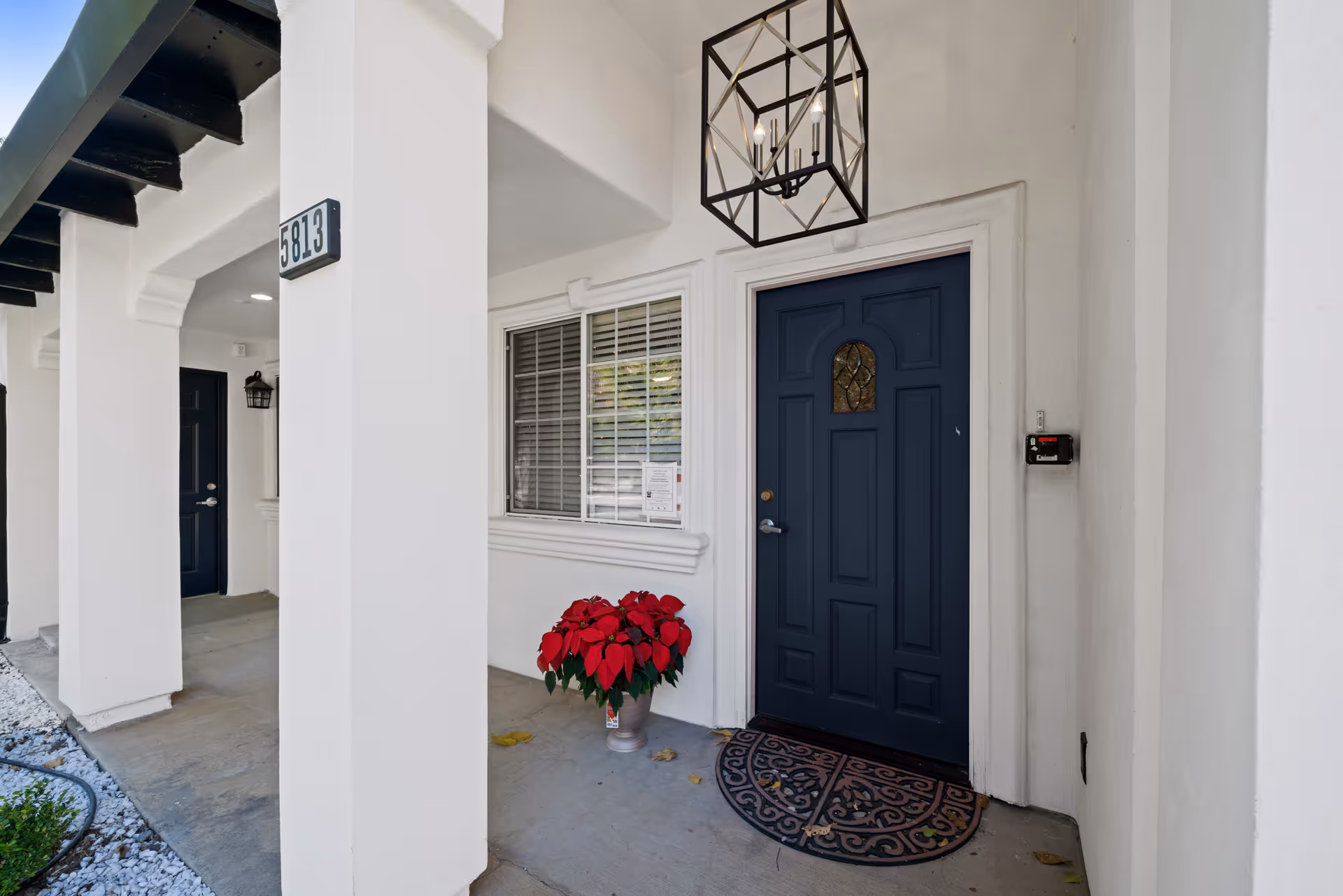 Entrance to a senior living unit with a dark blue door, a decorative doormat, a potted red poinsettia plant, white walls, and a window with blinds. The porch area has white columns and a modern black lantern-style light fixture hanging from the ceiling.