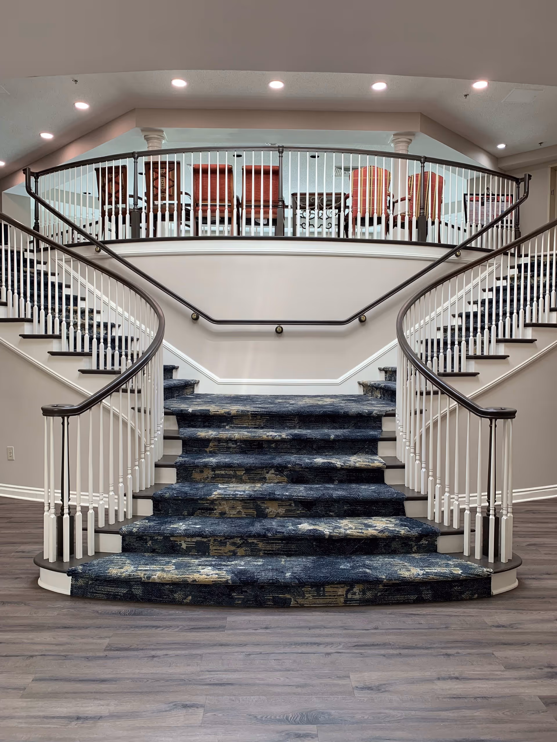A wide staircase with dark patterned carpet and white railings curves upward to a second-floor landing. The landing has a railing with white spindles and dark handrails, behind which several red and striped upholstered chairs are arranged. The ceiling above has recessed lighting, and the floor at the base of the stairs is a light wood laminate.