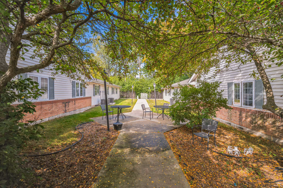Outdoor courtyard area with a concrete pathway leading between two single-story buildings with white siding and brick accents. There are several metal tables and chairs placed on the grass and concrete, surrounded by trees and bushes with green leaves. The sky is clear and blue.