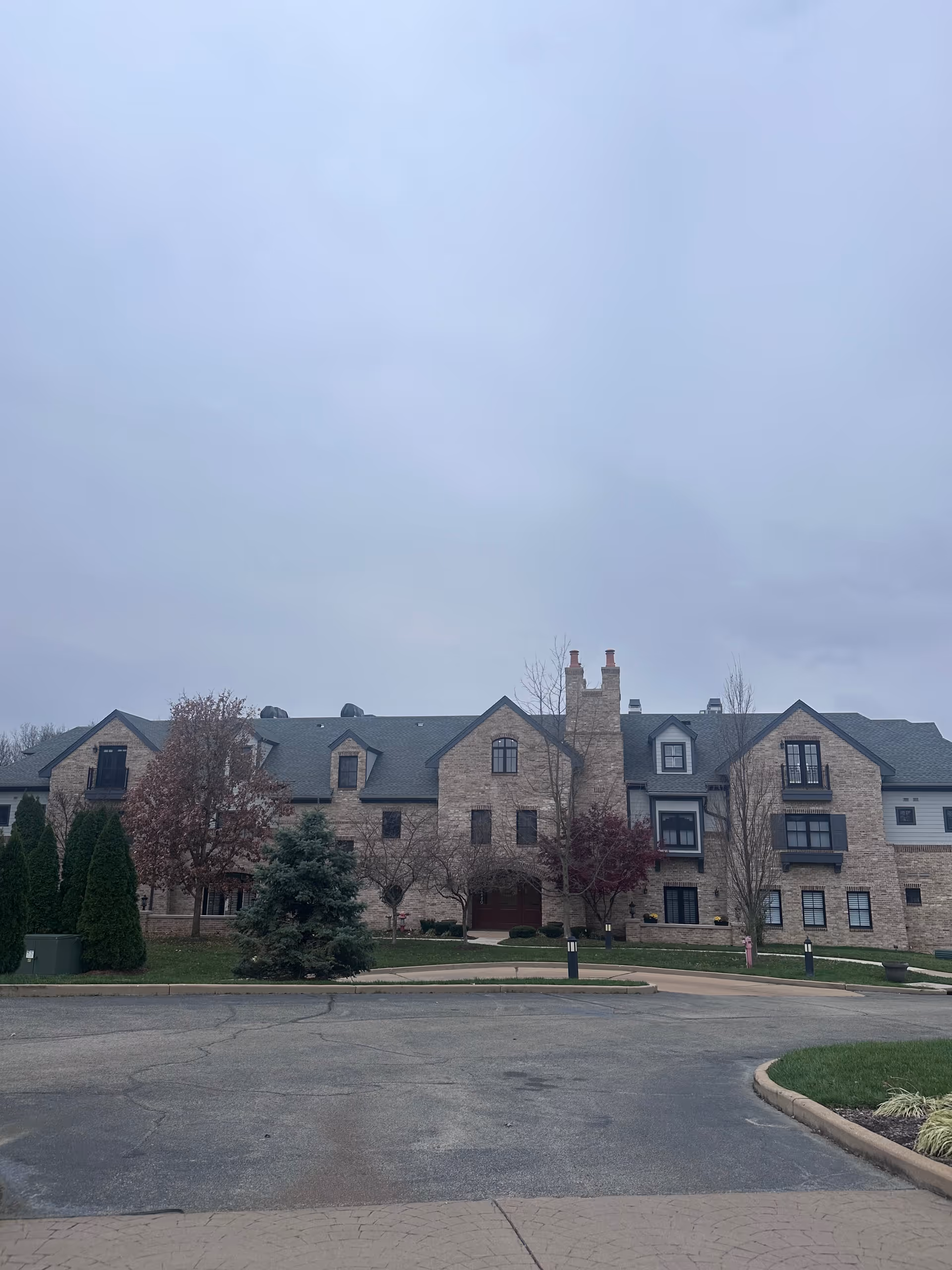 Stone-faced multi-story senior living building with dormer windows, landscaped trees and a circular driveway under an overcast sky.